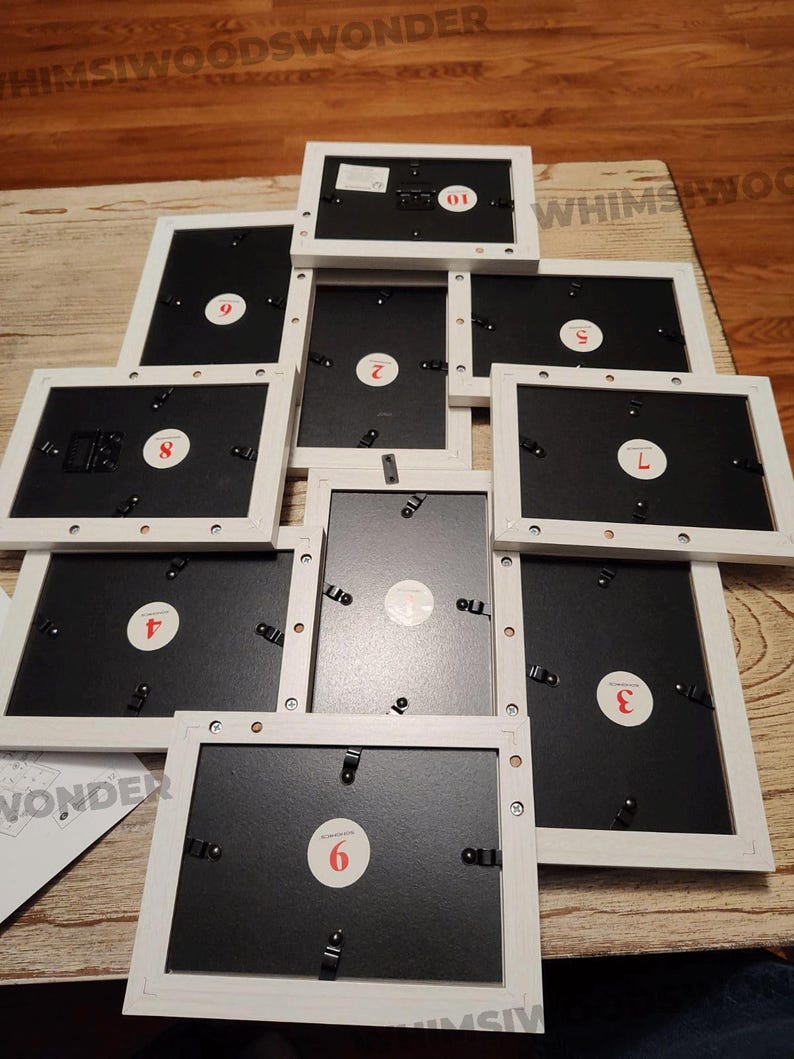 May include: Nine white picture frames with black backing and numbered labels from 1 to 9. The frames are stacked in a pyramid shape on a wooden surface.