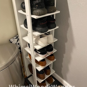 May include: White shoe rack with six shelves, holding a variety of shoes, including sneakers, sandals, and dress shoes. The rack is against a white wall and has the text "Whims Woods Wonder" on the top and bottom.