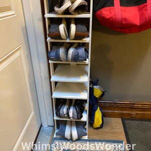 May include: White shoe rack with five shelves, holding pairs of shoes. The rack is against a white wall with a door to the left. A red and black tote bag hangs on the wall to the right of the rack. A blue and beige patterned rug is on the floor in front of the rack.