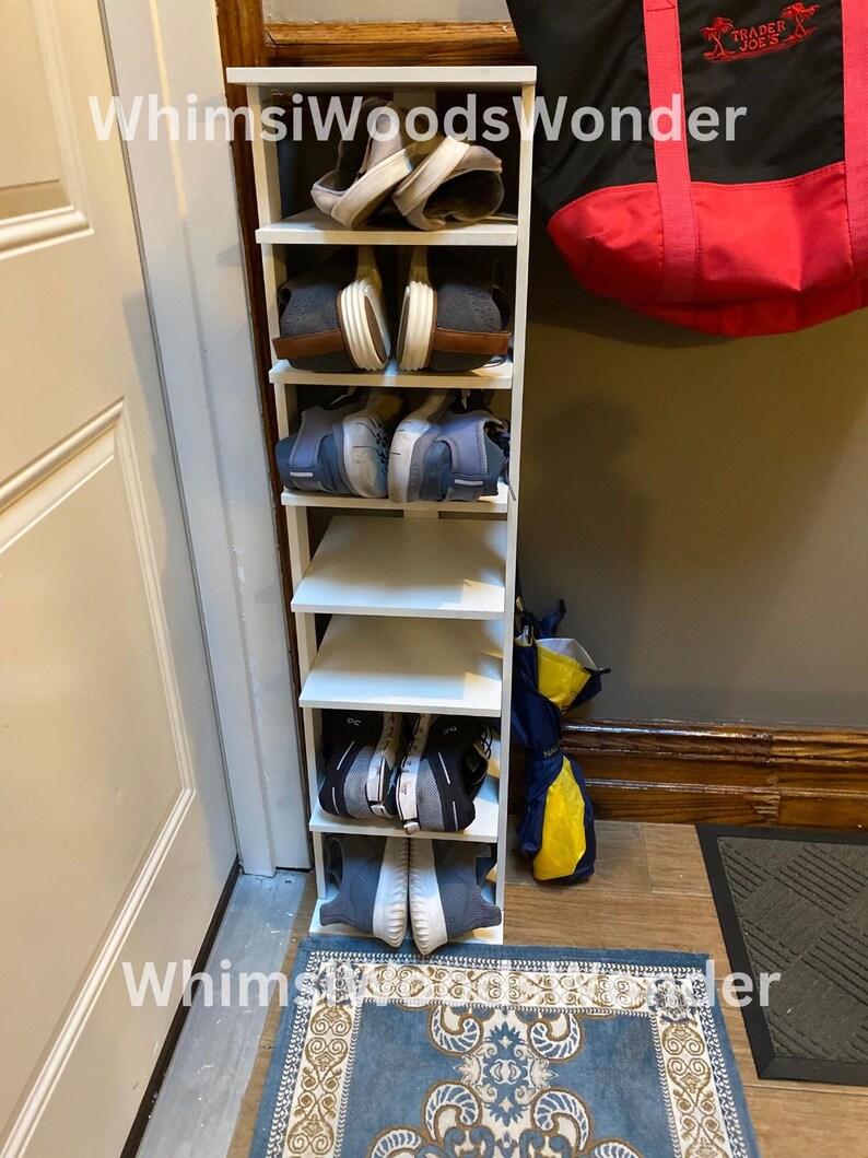 May include: White shoe rack with five shelves, holding pairs of shoes. The rack is against a white wall with a door to the left. A red and black tote bag hangs on the wall to the right of the rack. A blue and beige patterned rug is on the floor in front of the rack.