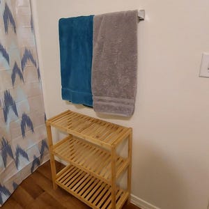 Puede incluir: Una estanter&iacute;a de bamb&uacute; de tres niveles con un acabado en madera clara. La estanter&iacute;a est&aacute; vac&iacute;a y est&aacute; sobre un suelo de madera. Dos toallas est&aacute;n colgadas en un toallero en la pared detr&aacute;s de la estanter&iacute;a. Una toalla es azul turquesa y la otra es gris.