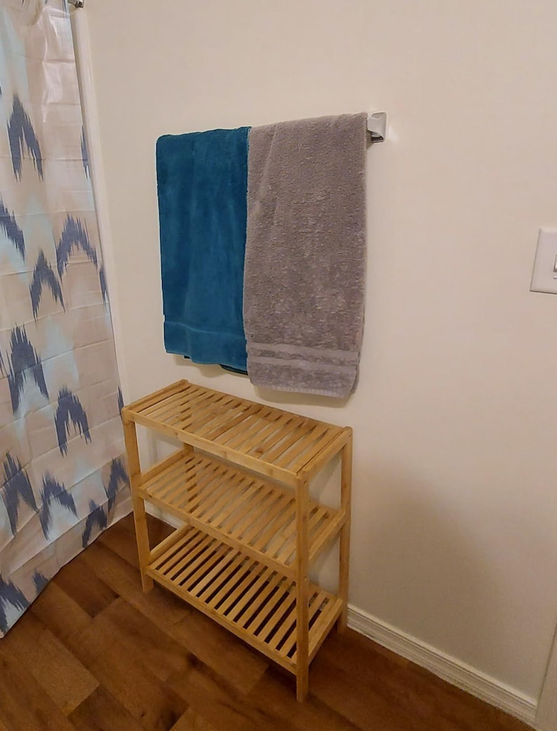 Puede incluir: Una estanter&iacute;a de bamb&uacute; de tres niveles con un acabado en madera clara. La estanter&iacute;a est&aacute; vac&iacute;a y est&aacute; sobre un suelo de madera. Dos toallas est&aacute;n colgadas en un toallero en la pared detr&aacute;s de la estanter&iacute;a. Una toalla es azul turquesa y la otra es gris.