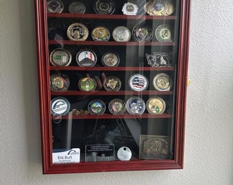 May include: A mahogany wood shadow box with a glass front displays a collection of round metal challenge coins. The shadow box has a black felt backing and is mounted on a white wall. A name card with the text "Eric Burt" is visible in the bottom right corner.