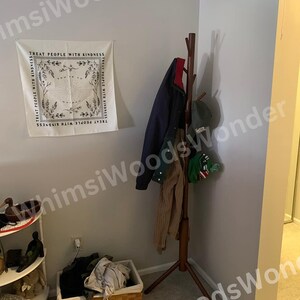 May include: A corner of a room with a wooden coat rack holding a navy jacket, a tan sweater, and a green hat. A white tapestry with the text "TREAT PEOPLE WITH KINDNESS" hangs on the wall. A wicker basket with clothes sits on the floor.