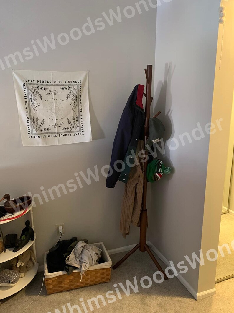 May include: A corner of a room with a wooden coat rack holding a navy jacket, a tan sweater, and a green hat. A white tapestry with the text "TREAT PEOPLE WITH KINDNESS" hangs on the wall. A wicker basket with clothes sits on the floor.