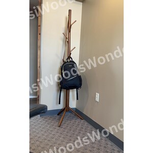 May include: A brown wooden coat rack with multiple branches for hanging items. A black backpack hangs from one of the branches. The rack stands on a three-legged base, and is placed against a beige wall.