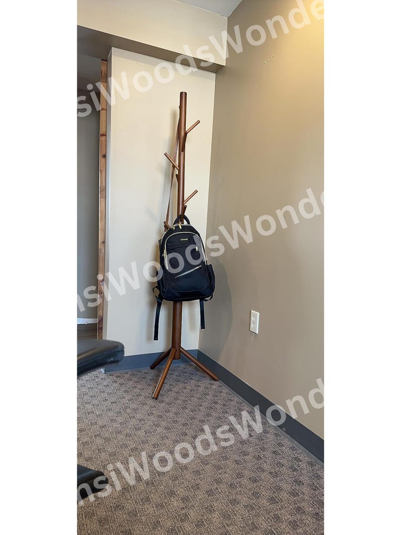 May include: A brown wooden coat rack with multiple branches for hanging items. A black backpack hangs from one of the branches. The rack stands on a three-legged base, and is placed against a beige wall.
