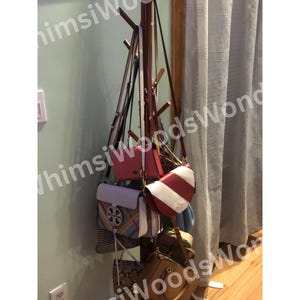 May include: A wooden coat rack displaying a variety of handbags. The handbags are in various colors, including red, white, brown, and blue. The rack is positioned against a light green wall and a white curtain.