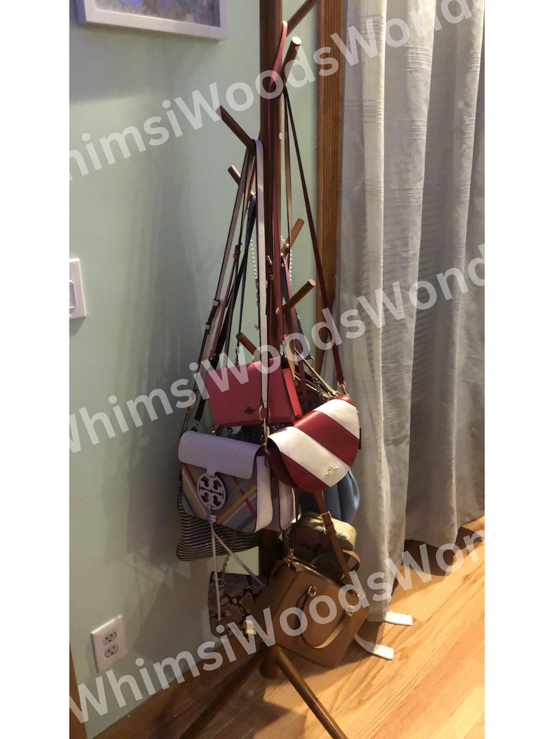 May include: A wooden coat rack displaying a variety of handbags. The handbags are in various colors, including red, white, brown, and blue. The rack is positioned against a light green wall and a white curtain.