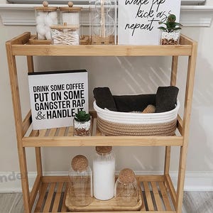Puede incluir: Una estanter&iacute;a de bamb&uacute; de tres niveles con un cartel blanco que dice "wake up kick ass repeat" y un cartel blanco y negro que dice "Drink some coffee put on some gangster rap & handle it". La estanter&iacute;a est&aacute; llena de varios art&iacute;culos, incluyendo bolas de algod&oacute;n, bastoncillos de algod&oacute;n, un frasco de vidrio con palitos, una peque&ntilde;a planta en maceta, una cesta tejida en blanco y negro y tres frascos de vidrio con tapas de corcho.