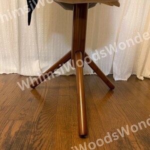 May include: A wooden, three-legged stool with a round top. The stool has a dark brown finish and is set against a white curtain background. The stool's legs are angled outwards for stability.