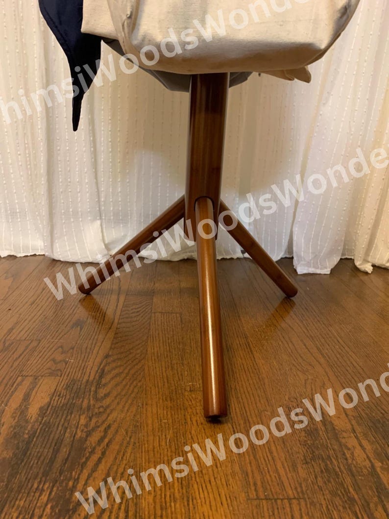 May include: A wooden, three-legged stool with a round top. The stool has a dark brown finish and is set against a white curtain background. The stool's legs are angled outwards for stability.