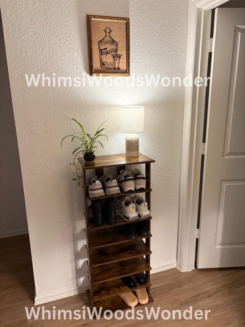 May include: A dark brown wooden shoe rack with six shelves, holding various shoes, including sneakers, boots, and sandals. A small table lamp with a white shade sits on top of the rack, and a framed picture of a tequila bottle hangs on the wall behind it.