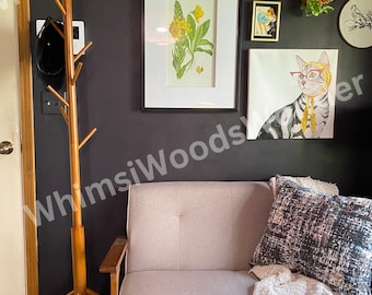 May include: A living room scene with a light gray sofa, throw pillows, and a cozy blanket. The wall features framed artwork, including a botanical print and a cat portrait. A wooden coat rack stands to the left.