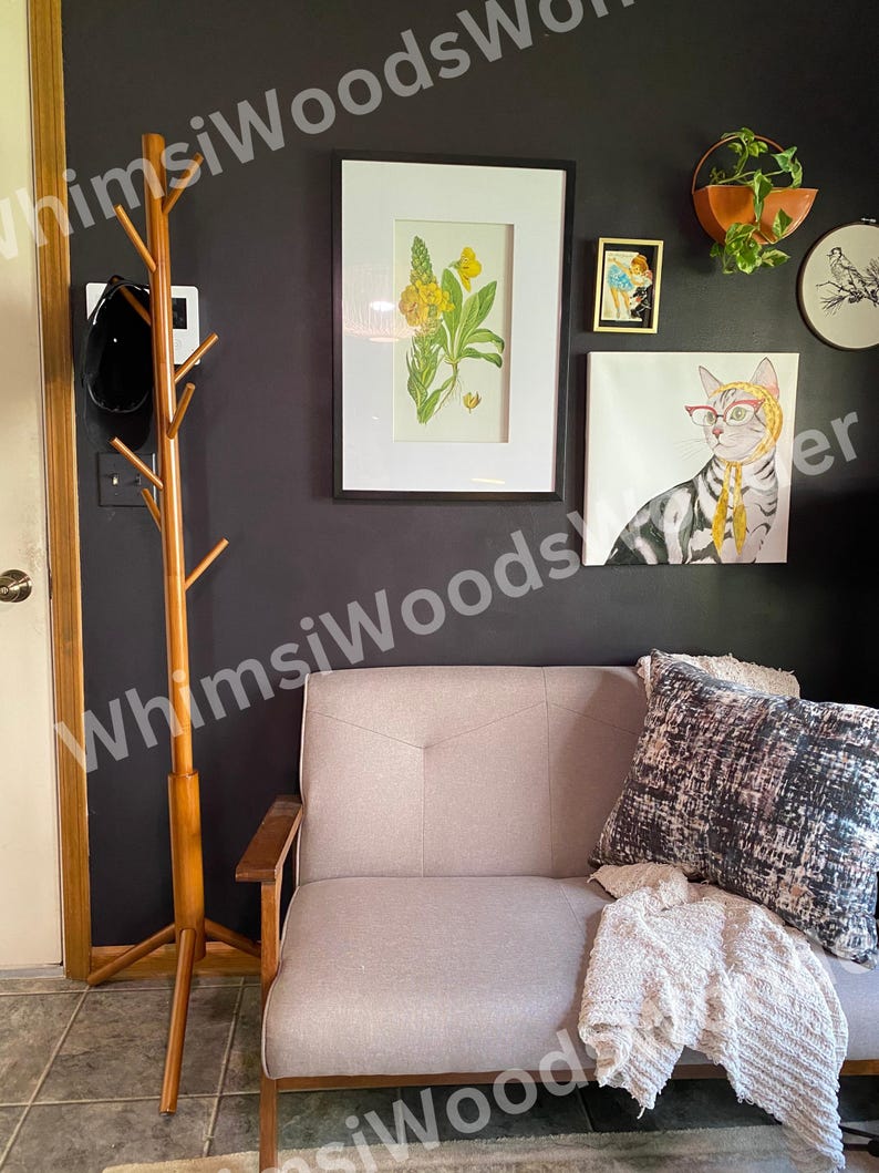 May include: A living room scene with a light gray sofa, throw pillows, and a cozy blanket. The wall features framed artwork, including a botanical print and a cat portrait. A wooden coat rack stands to the left.