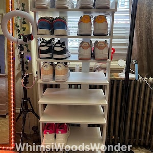 May include: White shoe rack with six shelves, filled with various pairs of sneakers, including Nike Air Force 1s, Nike Air Maxes, and other brands. The rack is against a white wall with a window in the background.