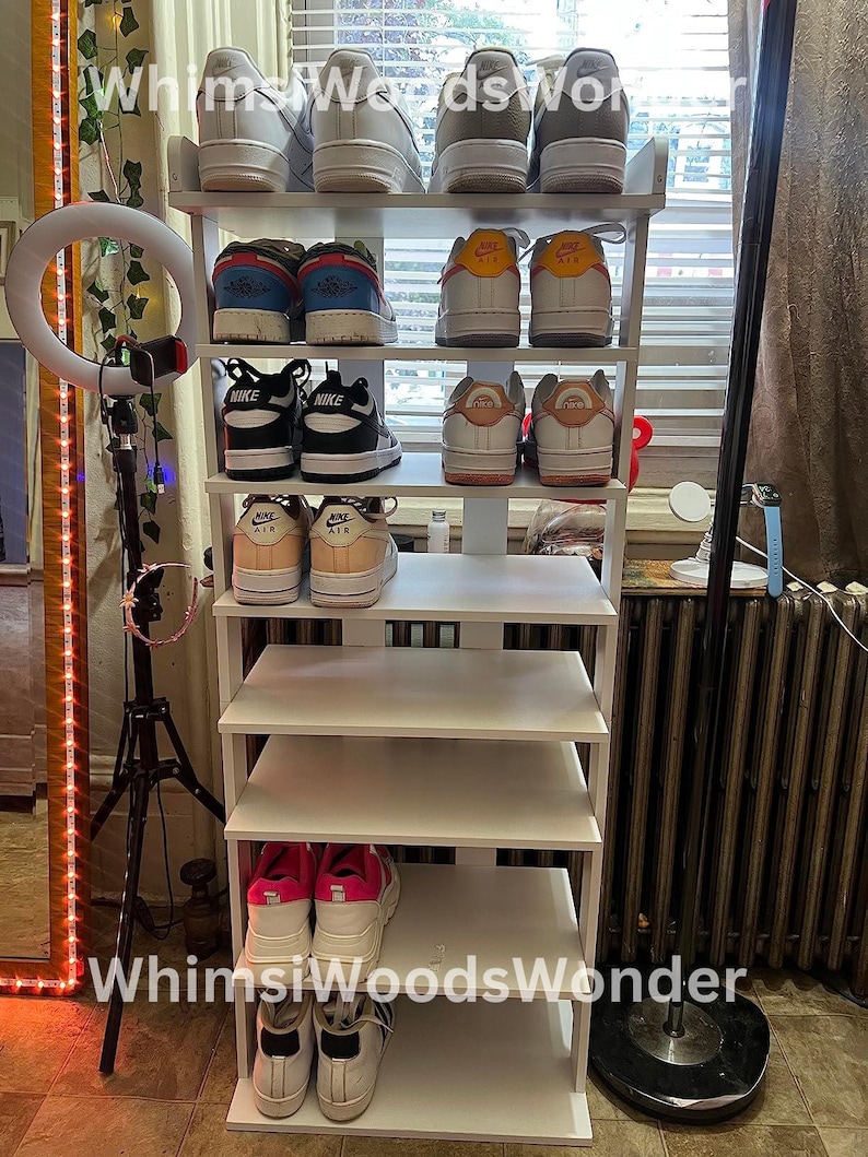 May include: White shoe rack with six shelves, filled with various pairs of sneakers, including Nike Air Force 1s, Nike Air Maxes, and other brands. The rack is against a white wall with a window in the background.