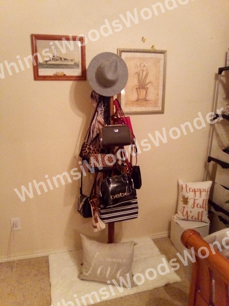 May include: A wooden coat rack displaying a gray fedora, various handbags, and a leopard print scarf. The rack is adorned with several purses in different styles and colors, including black, pink, and white. Two framed pictures hang on the wall.