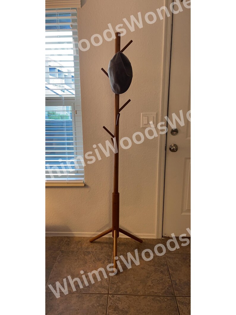 May include: A tall, wooden coat rack with a dark gray hat hanging from one of its pegs. The rack has a tripod base and multiple pegs for hanging coats or hats. The rack is made of a light brown wood.