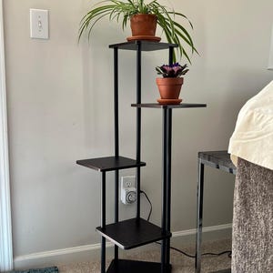 May include: A black metal plant stand with four shelves. The stand has a brown wood finish and is holding three potted plants. The plants are a spider plant, a purple flowering plant, and a green plant.