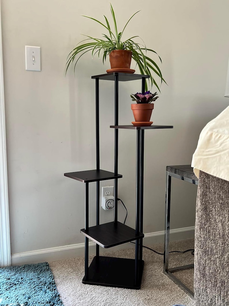 May include: A black metal plant stand with four shelves. The stand has a brown wood finish and is holding three potted plants. The plants are a spider plant, a purple flowering plant, and a green plant.