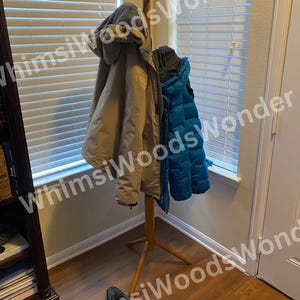 May include: A wooden coat rack with three coats hanging on it. The coats are tan and teal. Two pairs of black shoes are on the floor in front of the coat rack. The background includes a window with blinds and a door.