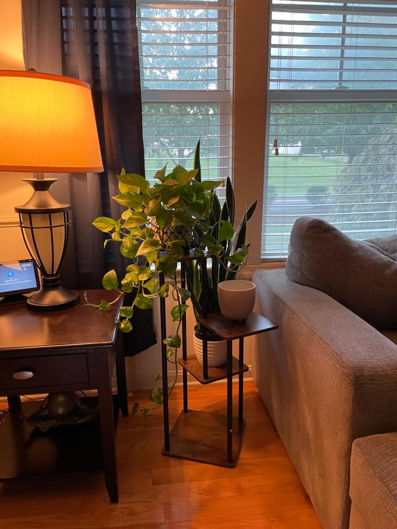 May include: A wooden plant stand with three shelves, holding two potted plants. The stand is in a living room setting, next to a couch and a side table with a lamp.