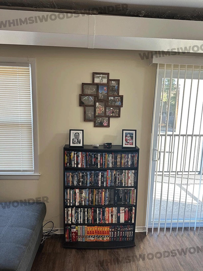 May include: A black DVD storage shelf with multiple rows of DVDs. The shelf is in a living room with a couch and a window with blinds. There is a picture frame with multiple photos on the wall above the shelf.