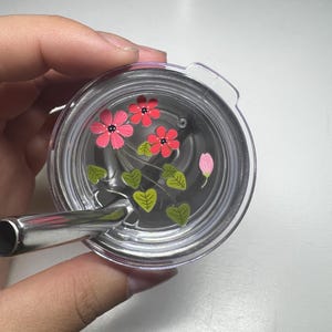 Puede incluir: Tapa de vaso transparente con pajita de acero inoxidable. La tapa presenta un dise&ntilde;o floral con flores rosas y rojas, hojas verdes y un capullo de flor rosa. La tapa del vaso est&aacute; sujeta por una mano.