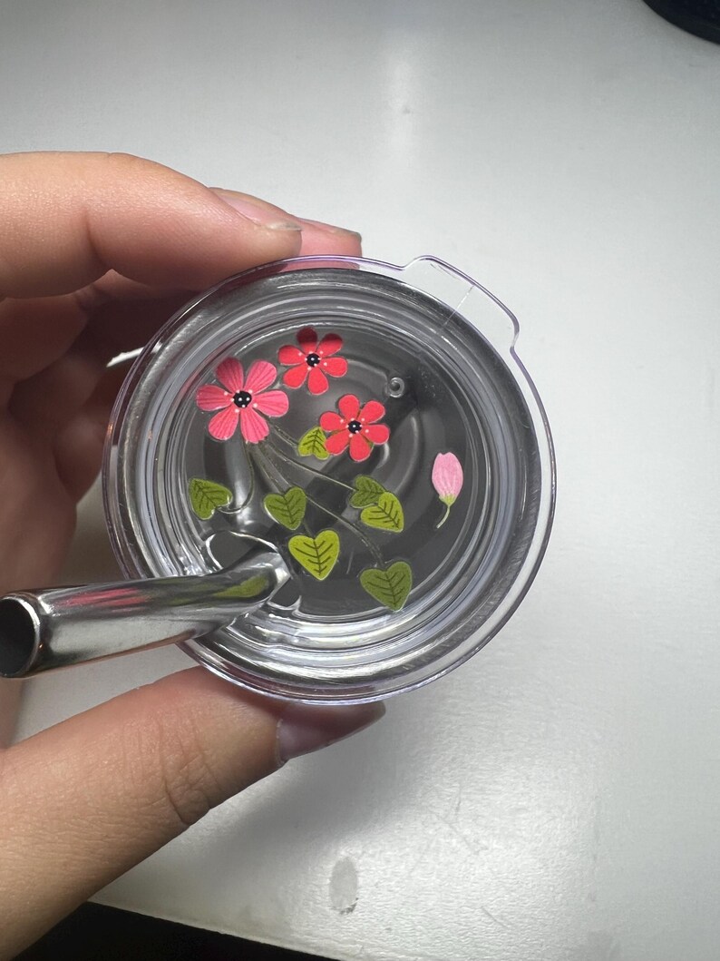 Puede incluir: Tapa de vaso transparente con pajita de acero inoxidable. La tapa presenta un dise&ntilde;o floral con flores rosas y rojas, hojas verdes y un capullo de flor rosa. La tapa del vaso est&aacute; sujeta por una mano.