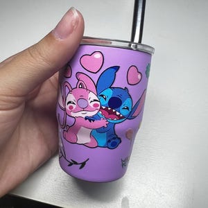 Puede incluir: Un vaso morado con una ilustraci&oacute;n de dibujos animados de Stitch y Angel abraz&aacute;ndose, decorado con corazones rosas y detalles florales. Tiene un borde plateado y una pajita negra. El dise&ntilde;o est&aacute; impreso en el exterior del vaso.
