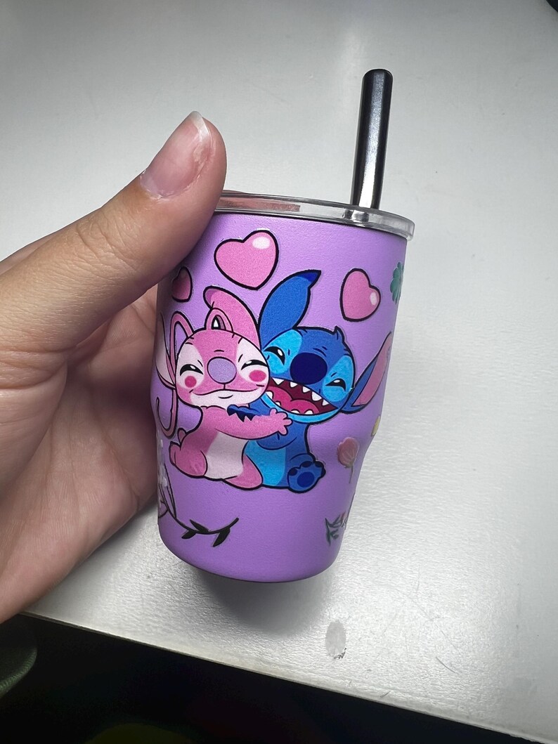 Puede incluir: Un vaso morado con una ilustraci&oacute;n de dibujos animados de Stitch y Angel abraz&aacute;ndose, decorado con corazones rosas y detalles florales. Tiene un borde plateado y una pajita negra. El dise&ntilde;o est&aacute; impreso en el exterior del vaso.