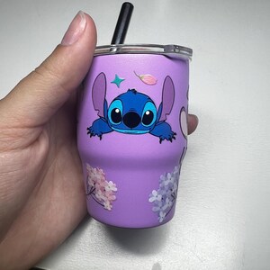 Puede incluir: Un vaso morado con un personaje de dibujos animados azul y una pajita negra. El vaso est&aacute; decorado con dise&ntilde;os florales y de estrellas. El vaso est&aacute; sostenido por una mano.