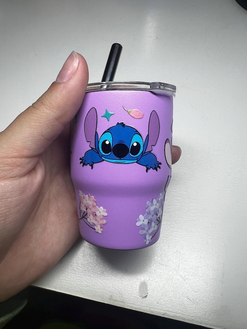 Puede incluir: Un vaso morado con un personaje de dibujos animados azul y una pajita negra. El vaso est&aacute; decorado con dise&ntilde;os florales y de estrellas. El vaso est&aacute; sostenido por una mano.