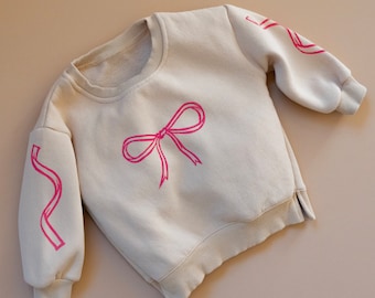 Sudadera beige de cuello redondo para bebés y niños con estampado de bloques de lazo y cinta rosa impresa a mano