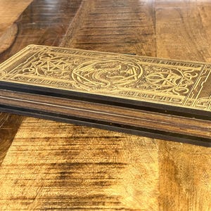 May include: A wooden box with a decorative, engraved design featuring a dragon and intricate patterns. The box is made of multiple layers of wood with a dark brown finish.