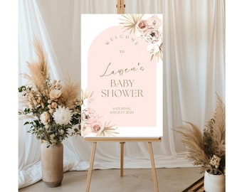 May include: Welcome to Lauren's Baby Shower sign with a pink background and gold lettering. The sign features a floral design with palm leaves and a white arch.