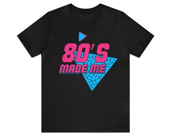 Puede incluir: Camiseta negra con un diseño gráfico rosa y azul. El gráfico presenta un triángulo con un botón de reproducción y el texto "80's MADE ME".