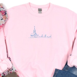 May include: A light pink crewneck sweatshirt featuring a blue embroidered line art illustration of a church and the text It is well with my soul, paired with a floral scarf, blue denim jeans, and taupe ankle boots with block heels.