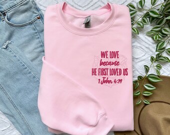 Könnte beinhalten: Rosafarbener Pullover mit einem Blumendesign und dem Text "We love because he first loved us 1 John 4:19".
