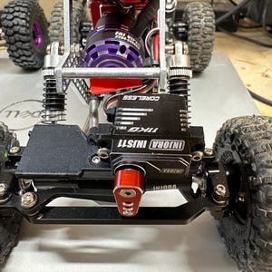 以下が含まれることがあります： 黒と赤のラジコンカーのシャーシのクローズアップ。黒と銀のモーターと黒と銀のサーボを搭載。車は、ノブタイヤを装着した黒いホイールを備えています。