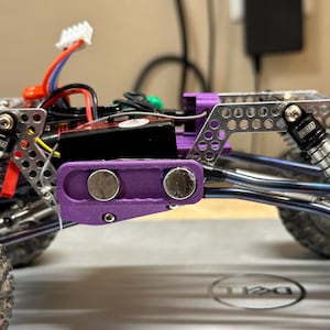 May include: A close-up of a silver and purple miniature off-road vehicle chassis with two shock absorbers. The chassis has a purple plastic body with two round magnets. The vehicle is on a gray surface.