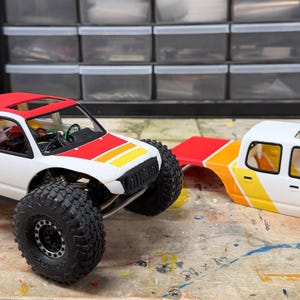 以下が含まれることがあります： 黒い車輪と黄とオレンジのストライプデザインの、白と赤のおもちゃのトラック。トラックは赤い屋根をしており、お揃いのトラックキャブの隣にあります。トラックはマルチカラーの表面にあります。