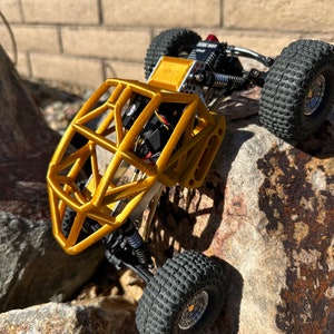 May include: A gold-colored, miniature, off-road vehicle with a roll cage, black tires, and a silver chassis. The vehicle is on a rocky surface.