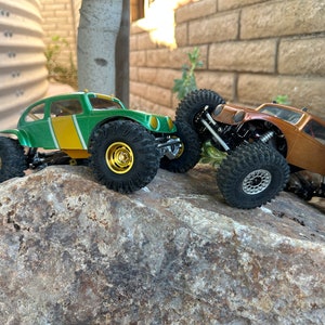May include: Two green and brown toy off-road vehicles with large tires are on a rock. The green vehicle has a yellow stripe on the side. The brown vehicle has a silver rim on the front tire.