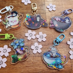 Peut inclure: Cinq porte-clés irisés représentant des personnages d'anime dans différentes tasses à thé et bols. Chaque porte-clés présente un personnage différent et une tasse ou un bol différent. Les porte-clés sont sur une surface en bois avec des décorations en forme de fleurs roses.