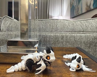 Pode incluir: Duas figuras de dragão branco e preto com olhos laranja são exibidas em uma superfície de madeira. Os dragões têm um design esquelético, com corpos articulados e detalhes. Um está em pé, o outro está deitado.