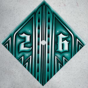 May include: A teal and white airbrushed design on a diamond-shaped surface. The design features the numbers "2" and "6" in a stylized font, with vertical lines and star-like accents. The background is a speckled gray.