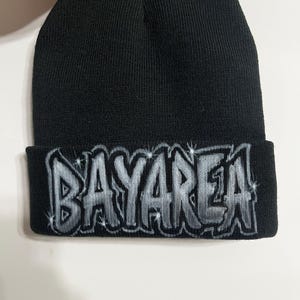 May include: Black knit beanie with the word "BAYAREA" in a silver and gray graffiti-style design across the folded brim. The beanie is made of a soft, ribbed knit material.
