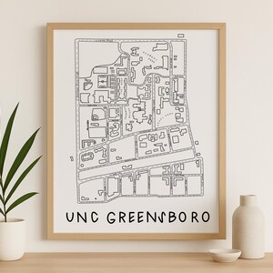 Op de afbeelding: Een ingelijste zwart-wit kaartafdruk van UNC Greensboro. De kaart toont de campusindeling met straten, gebouwen en groene ruimtes. De woorden "UNC GREENSBORO" zijn gedrukt in een vetgedrukte, schreefloze letter onderaan de afdruk. De lijst heeft een licht houten kleur.